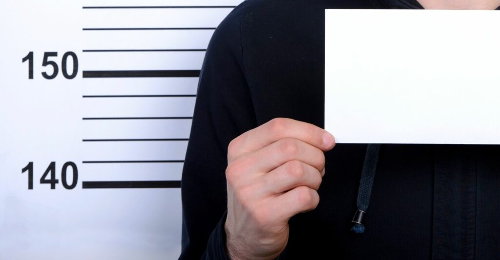 a shoulder to chest shot of a person's body (only their right hand side of the body) holding a blank white piece of paper in front of a background with height measurement lines displaying the numbers 140 and 150, insinuating a mug shot.