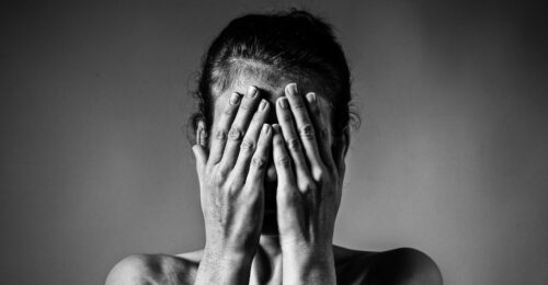 A black and white image of a women from her shoulders upward, with both hands covering her entire face.