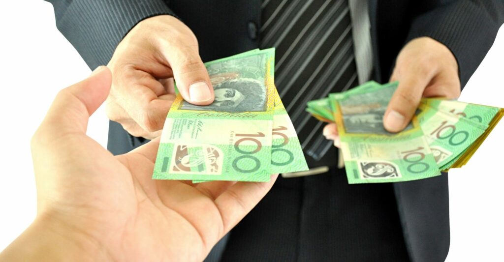 A photo showing the hands of two people standing opposite each other from one person's point of view. The person standing opposite is handing two one hundred Australian dollar notes with their right hand, and holding a stack of one hundred and fifty Australian dollar notes in their left hand.