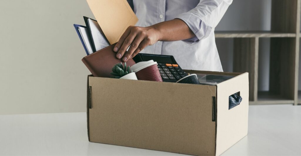 A photograph of a person's torso packing away their belongings into a cardboard box that is sitting on a table. Insinuating a person packing away their things from a workplace.