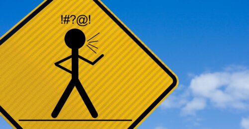 A close up of a yellow diamond road sign with a black human stick figure in the middle with one hand on its hip and the other raised in front. On top of the stick figure are the black characters "!#?@!" which is insinuating swearing.