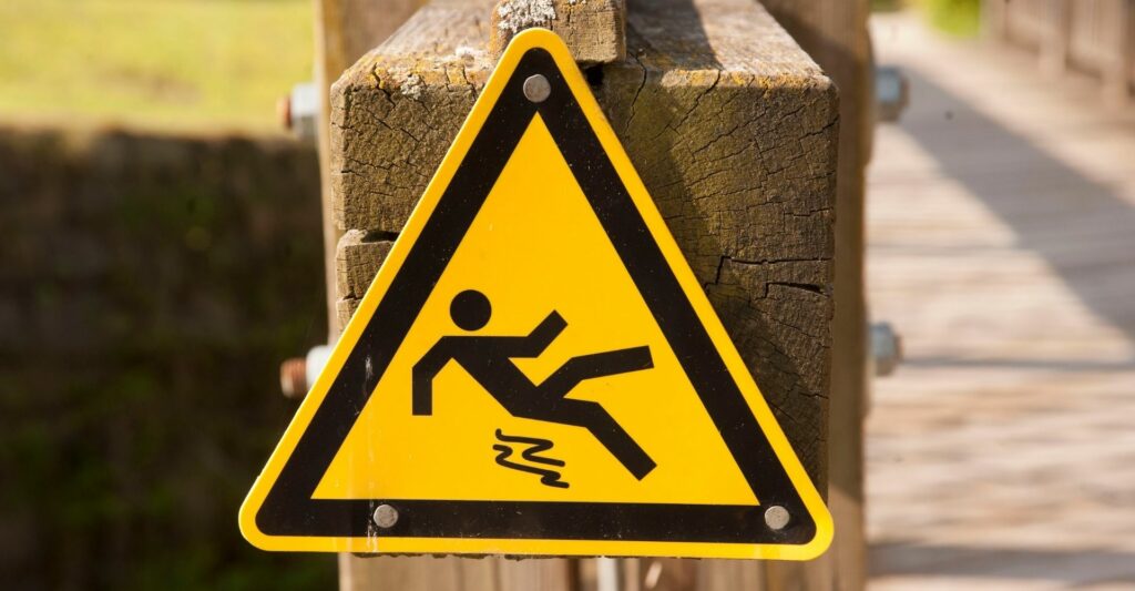 a yellow triangle sign nailed onto a wooden post. The sign shows a stick figure slipping on something and is about to fall down.