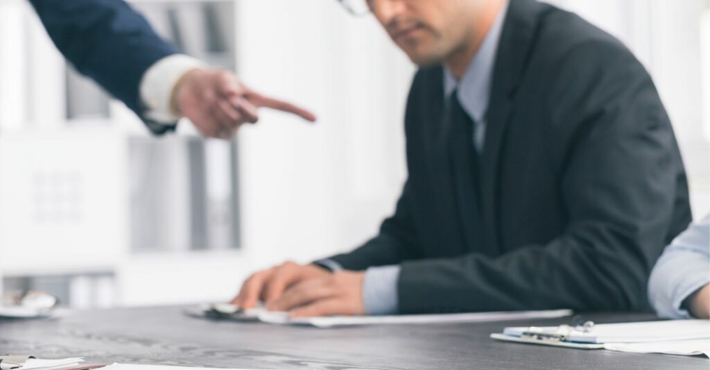 A blurry image of someone wearing a suit and glasses sitting at a meeting table and someone else on their right hand side pointing their index finger at them.