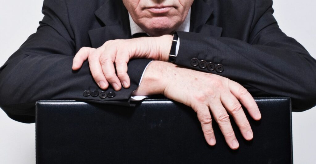 A photo of a man wearing a suit leaning his forearms on a black briefcase. He has a stern expression.