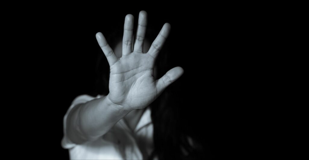 A half shot in black and white of a person extending their arms and showing the palm of their right hand, which covers their entire face. The motion is insinuating pushing something away or saying 'no'.