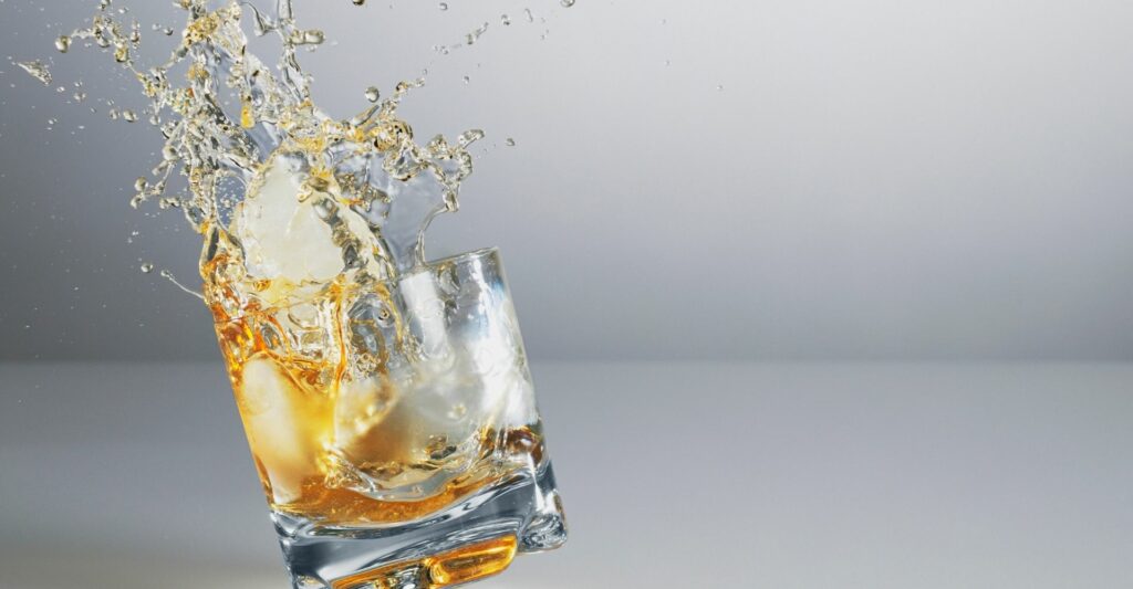 An in-action shot of a short glass with some alcohol (similar to whiskey or rum) in the middle of being knocked and the alcohol is splashing out of the glass.