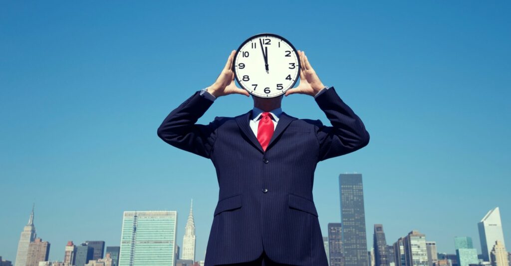 A suited man standing in front of a city skyline in the day time, holding an analogue clock with both hands and putting it in front of his face.