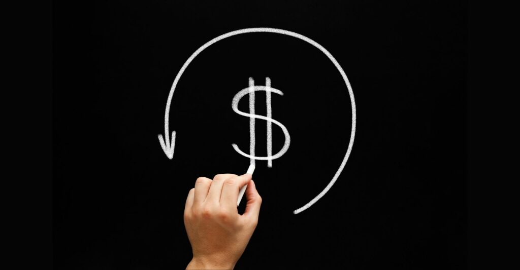 An image if someone's left hand holding a chalk and drawing a dollar sign in the middle of the chalkboard - on the outside of the dollar sign is an arrow going in a circular motion from the right hand side to the left.