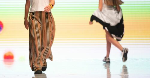 A lower half body shot of two children on the runway. The left child is walking towards the front wear brown baggy pants and a white t-shirt, and the child on the right is walking away from the frame wearing a sports jacket and sneakers.