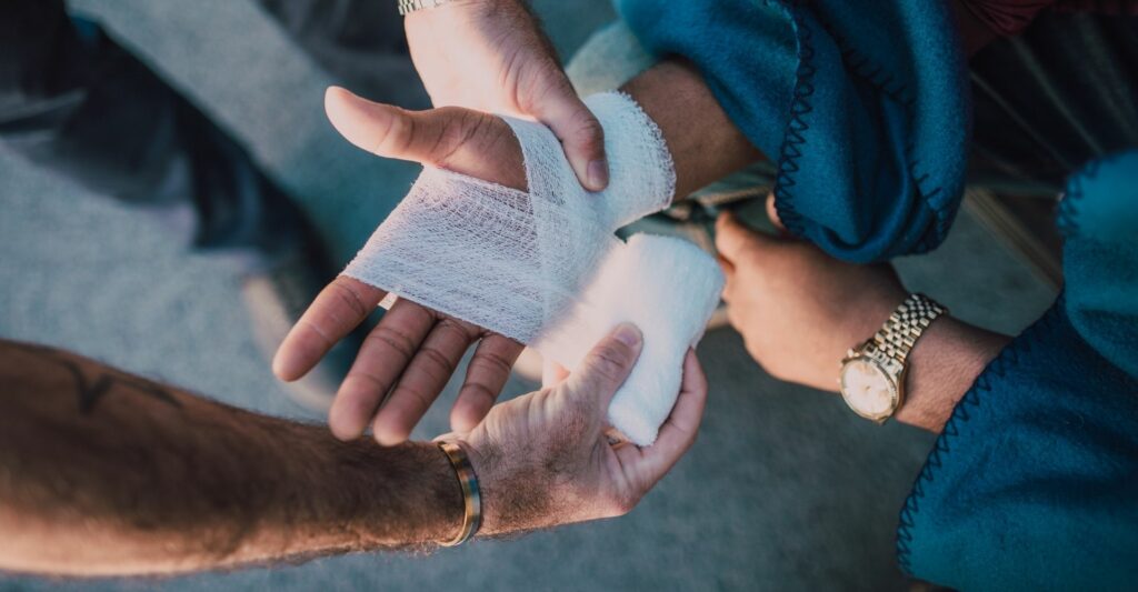 A photo of two people's hands. A person's right hand is being bandaged with a white bandage by another person to heal an injury.