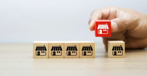 an image of a hand placing a red cube block onto the table in the line of five other wooden cube blocks. there is a white store front graphic printed on the front of these blocks.