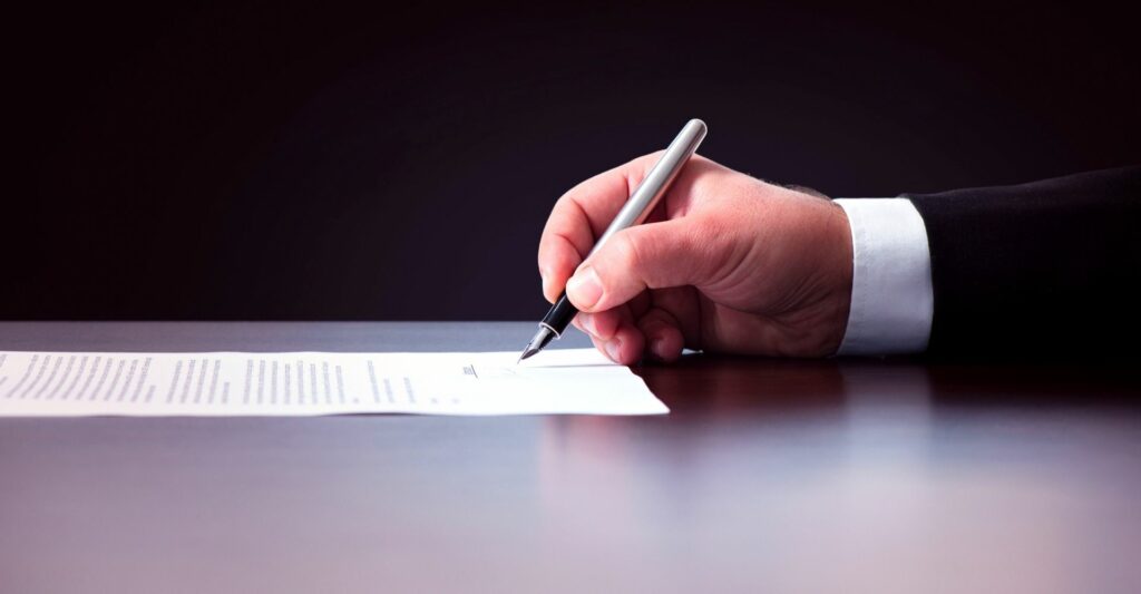 A side photo of someone's right hand holding a fountain pen and signing a piece of paper.