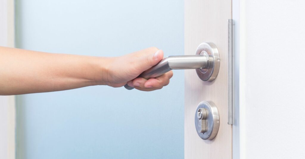 A close up photo of a person's left hand opening up a door by turning a silver door handle.