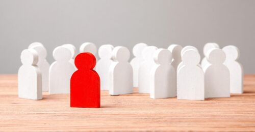 a photo of a group of white human shaped blocks standing on a table, and a red block stands in front.