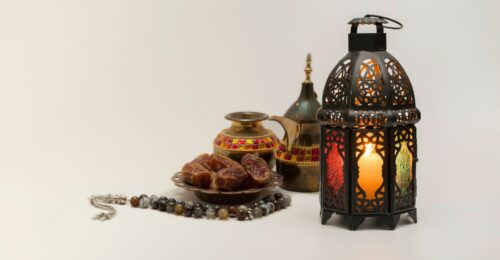 A photo of a bronze lit up lantern, and some utensils of a middle east style, with some dates sitting in a brass bowl.