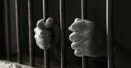 A black and white photo of two hands holding onto jail bars.