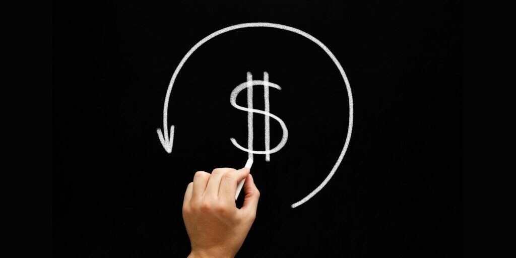 An image if someone's left hand holding a chalk and drawing a dollar sign in the middle of the chalkboard - on the outside of the dollar sign is an arrow going in a circular motion from the right hand side to the left.