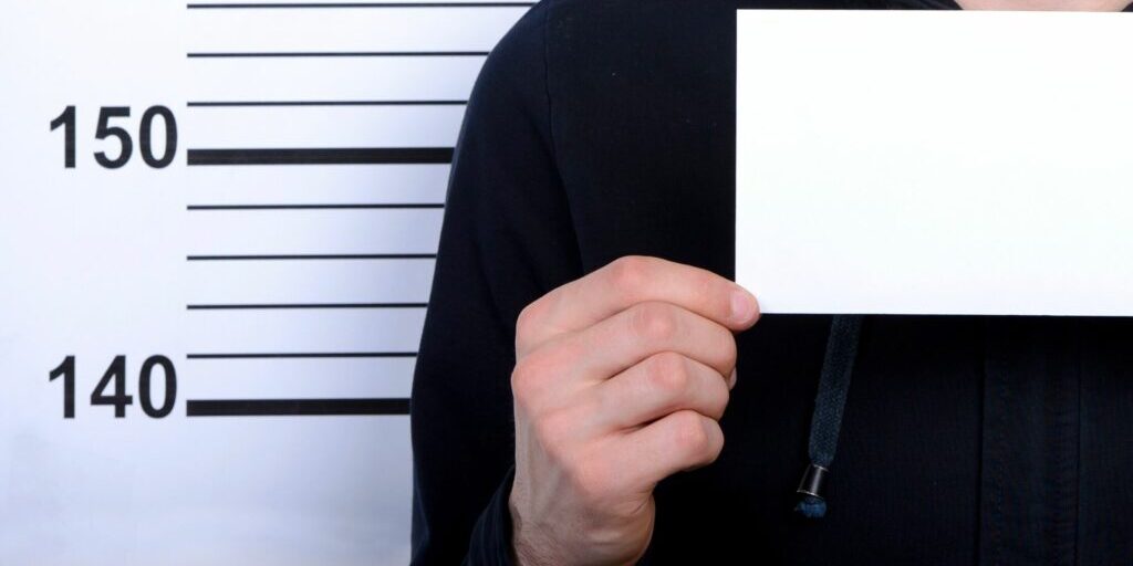 a shoulder to chest shot of a person's body (only their right hand side of the body) holding a blank white piece of paper in front of a background with height measurement lines displaying the numbers 140 and 150, insinuating a mug shot.