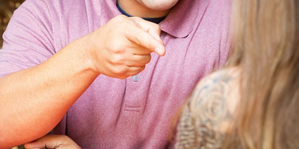 An torso shot of a man wearing a punk polo shirt pointing his index finger at a women. The woman's back of shoulder and parts of the back of her head can be seen on the right hand side of the image - slightly blurred.