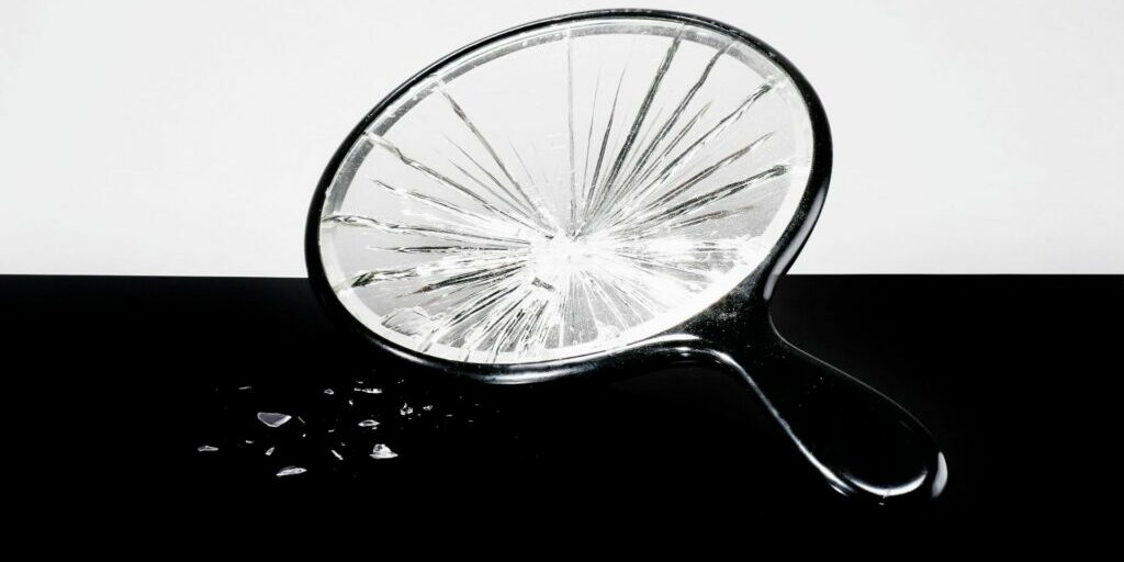 a broken black magnifying glass with small shards of glass to the left of the photo, The background consists of two landscape rectangles, the top is an off white, and the bottom is black.