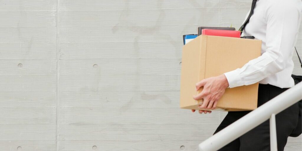 A torso shot of a person walking down the stairs wearing work clothes and holding a cardboard box filled with belongings.