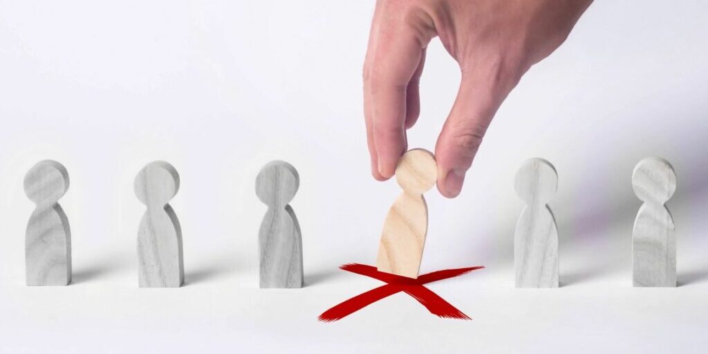 An image of someone's right hand picking up a wooden block in a shape of a simple person that was standing on a red 'X'. The wooden block is in the middle of a line of other grey blocks of the same shape.