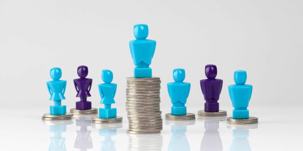 A small blue pawn statue standing on top of a large stack of coins in the middle of the image, and behind it are six other blue and purple pawns standing on top of a lower stack of coins.