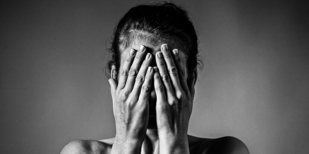 A black and white image of a women from her shoulders upward, with both hands covering her entire face.