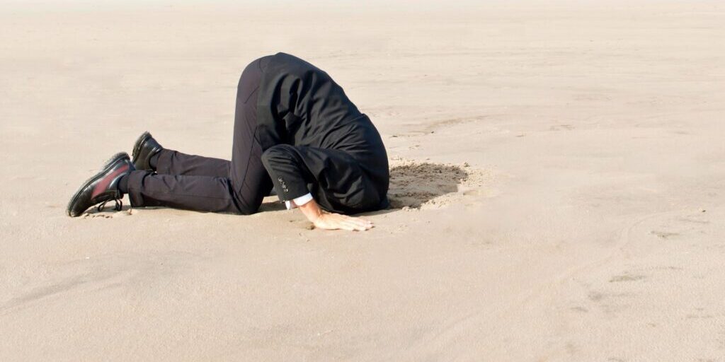 An image of a man in a black suit and black leather shoes with his entire hand in the sand as the rest of his body is kneeling on the sand.