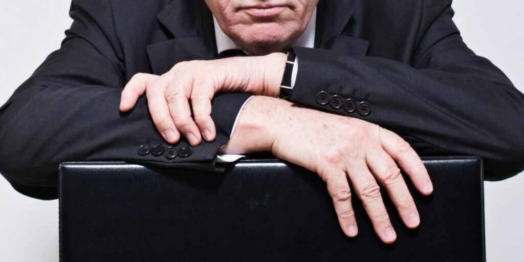 A photo of a man wearing a suit leaning his forearms on a black briefcase. He has a stern expression.