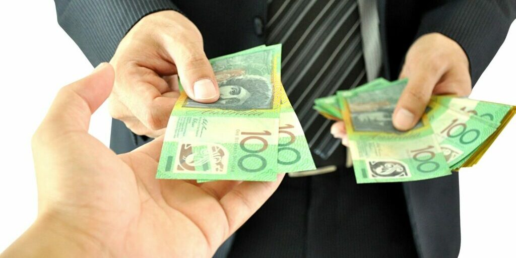 A photo showing the hands of two people standing opposite each other from one person's point of view. The person standing opposite is handing two one hundred Australian dollar notes with their right hand, and holding a stack of one hundred and fifty Australian dollar notes in their left hand.