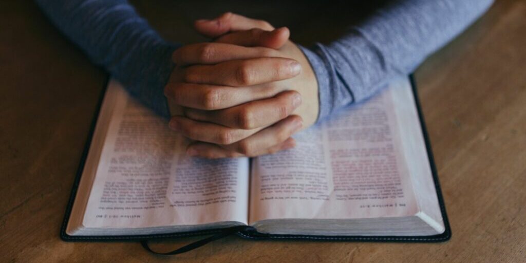 A shot of a person's hands with their fingers clasped together laid in the middle of an open book.