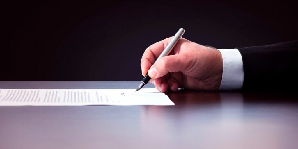 A side photo of someone's right hand holding a fountain pen and signing a piece of paper.