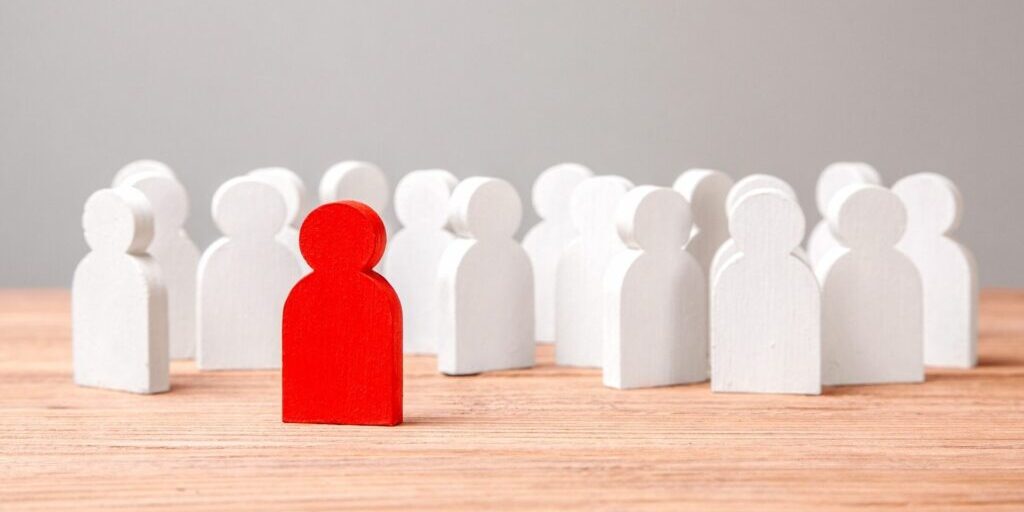 a photo of a group of white human shaped blocks standing on a table, and a red block stands in front.