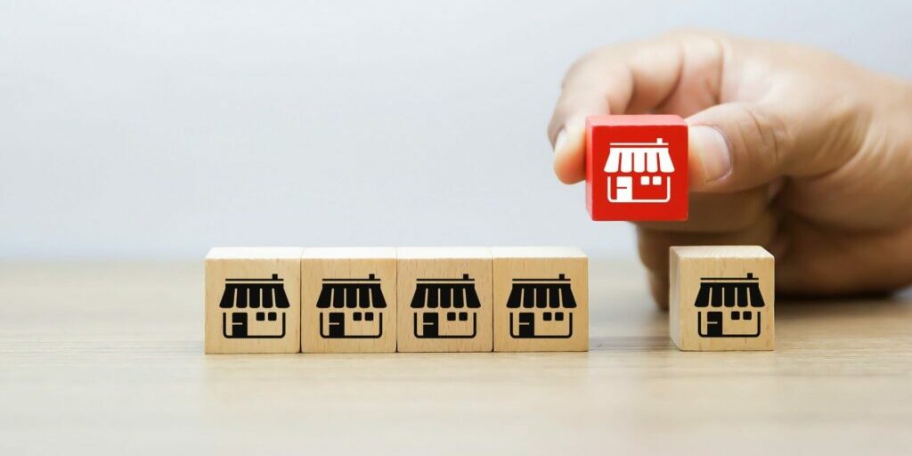 an image of a hand placing a red cube block onto the table in the line of five other wooden cube blocks. there is a white store front graphic printed on the front of these blocks.