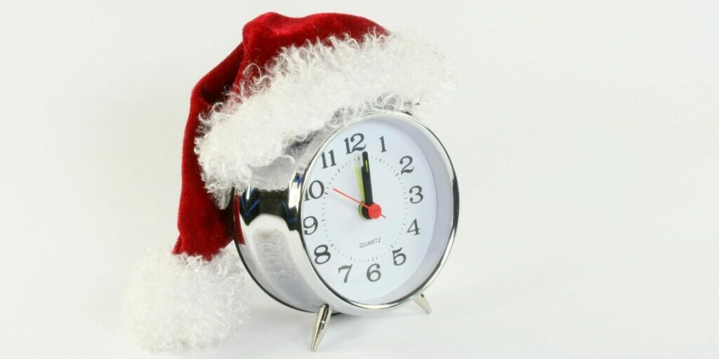 A photograph of a silver alarm clock with a christmas santa hat sitting on the top of it.