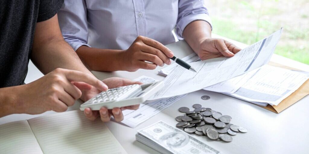 A photo of two people sitting at a desk and only their hands and forearms can be seen. The person on the left is holding a document in his left hand and a pen in his right hand and is using the pen to point at the document. The person on the right is holding a calculator and inputting a function. On the table are a couple of documents, a cheque, a stack of coins, a pile of notes and an open, blank, lined notebook.