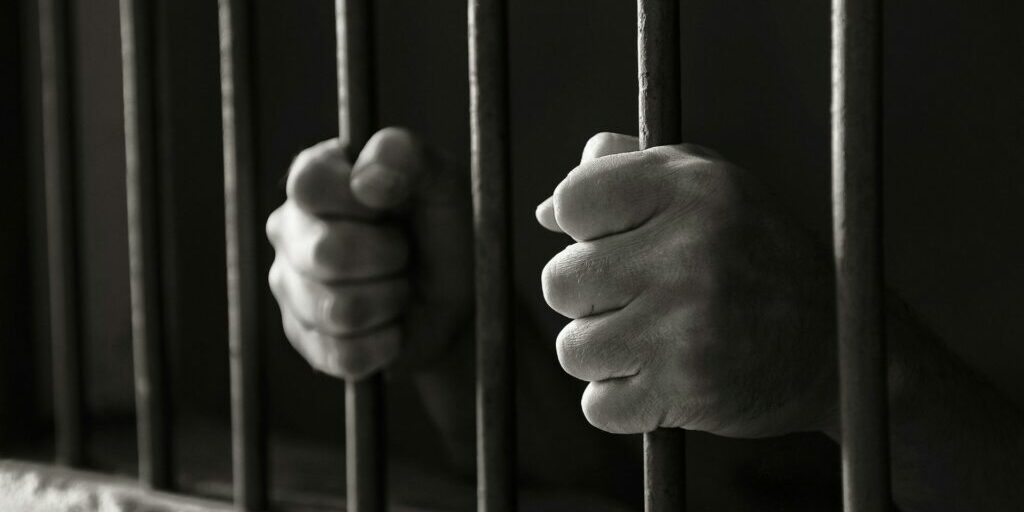 A black and white photo of two hands holding onto jail bars.