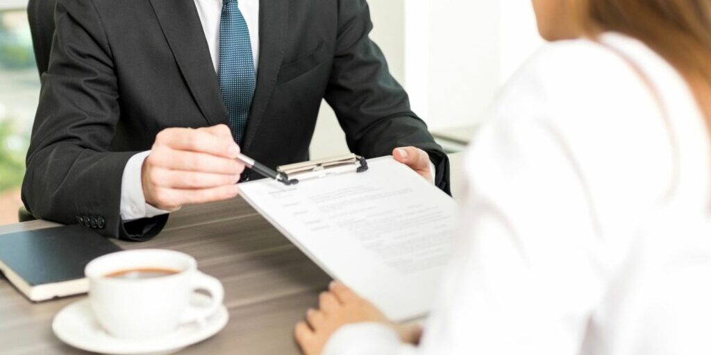 a photo showing a man in a suit holding a clipboard (with a document in it) and pen and showing it to a woman sitting opposite him. The pen he is holding is pointing to a section in the document.