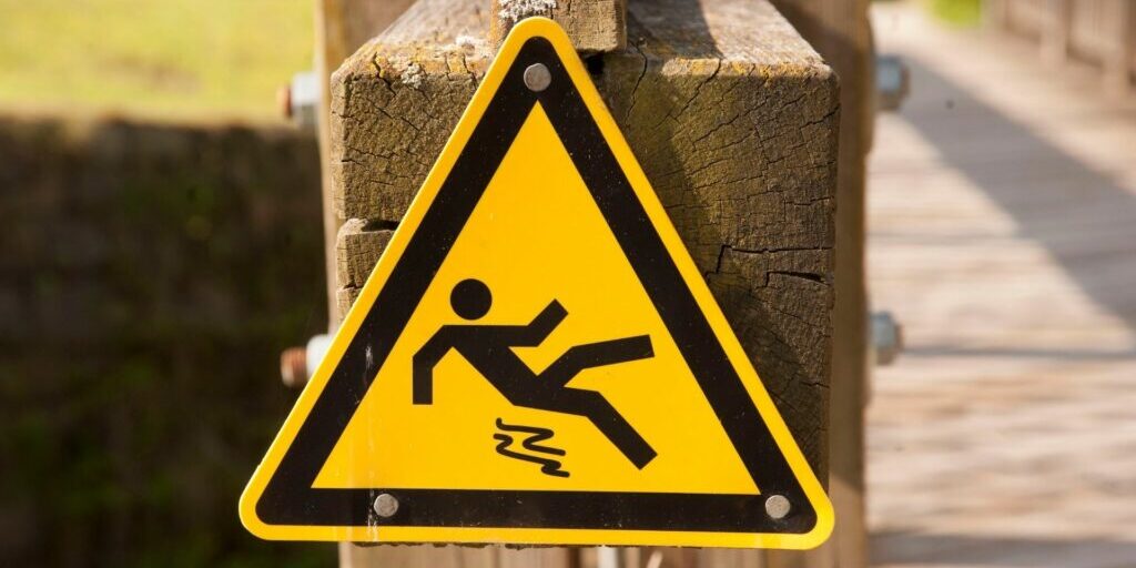 a yellow triangle sign nailed onto a wooden post. The sign shows a stick figure slipping on something and is about to fall down.