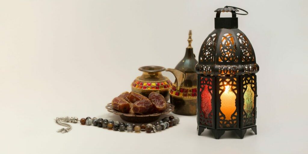 A photo of a bronze lit up lantern, and some utensils of a middle east style, with some dates sitting in a brass bowl.