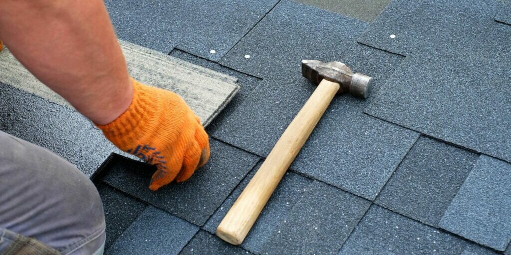 A photograph of a person placing a tile down on top of other tiles. Only his right hand forearm and parts of his knee can be seen. To his right is a hammer that sits in the middle of the image.