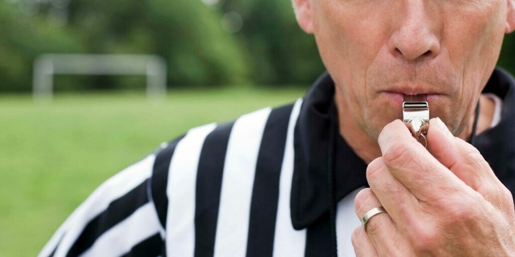 A close up of a referee's face as he blows a metal whistle with his left hand. The referee is wearing a black and white striped shirt and is standing on a grass sports field with a white goal post.