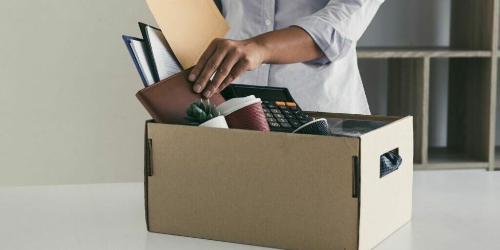 A photograph of a person's torso packing away their belongings into a cardboard box that is sitting on a table. Insinuating a person packing away their things from a workplace.
