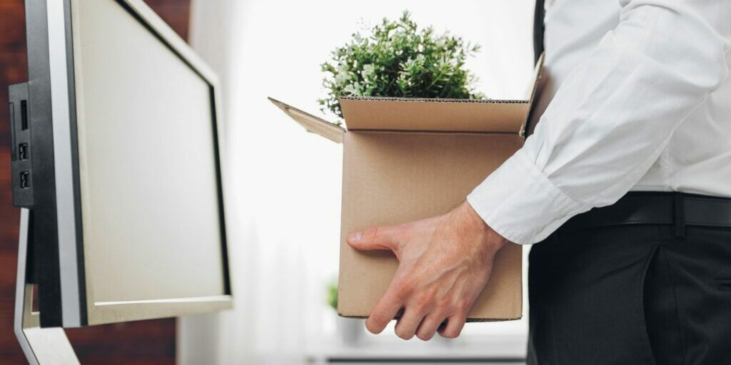 a side shot of a person's torso while they are holding a cardboard box with a plant sticking out, as he stands in front of a desktop monitor.