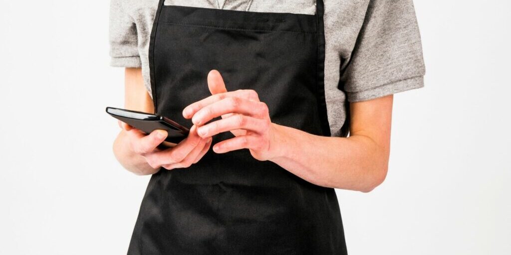 A photo of worker's torso who is wearing a black apron and holding a dark brown notebook - similar to the ones waitresses use to write down menu orders.