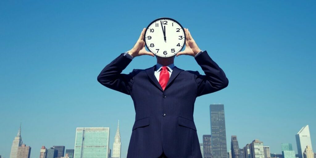 A suited man standing in front of a city skyline in the day time, holding an analogue clock with both hands and putting it in front of his face.