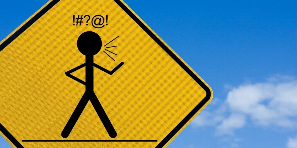 A close up of a yellow diamond road sign with a black human stick figure in the middle with one hand on its hip and the other raised in front. On top of the stick figure are the black characters "!#?@!" which is insinuating swearing.
