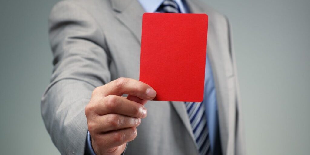 A torso shot of a man in a grey blazer, light blue collared dress shirt and a blue stripy tie holding up a red rectangle card.