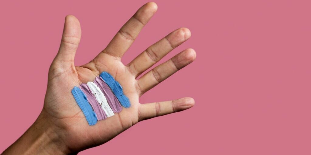 A photo of the inside of someone's left hand with the fingers spread, and on the palm is painted with 5 painted lines in the order (from left to right) of blue, pink, white, pink and blue. These painted links make up a sort of square right in the middle of the palm. The background of the hand is a blush pink.